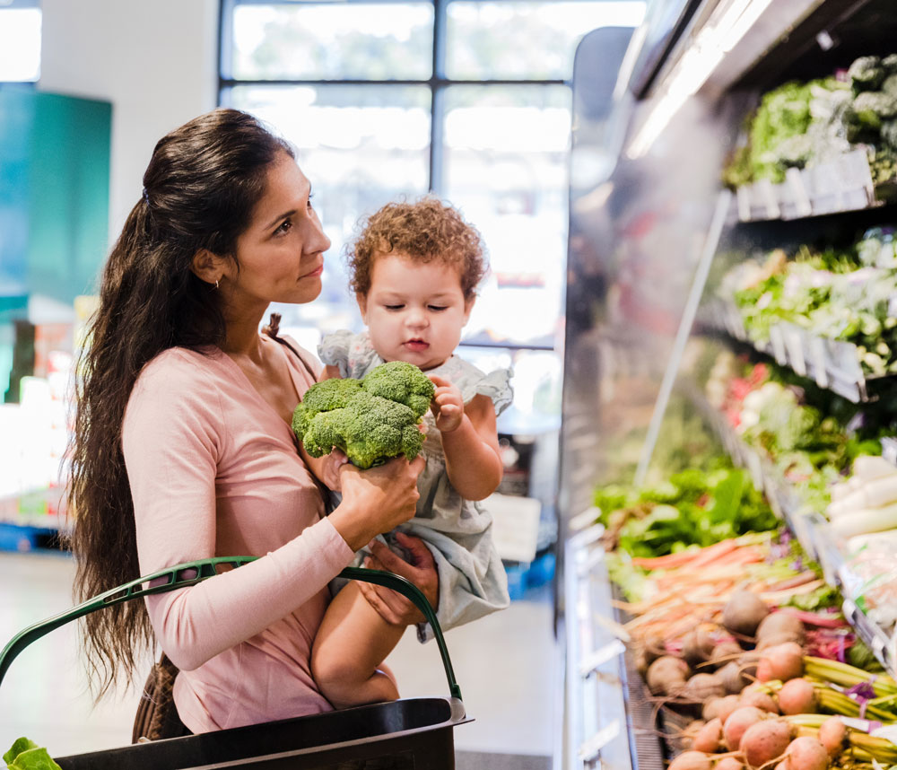 Mother holding baby while grocery shopping for vegetables